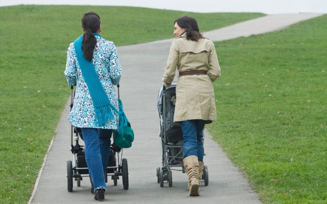 Two women walk side by side on a paved path in a grassy park, each pushing a stroller. The sky is overcast, and the path curves gently uphill in the distance.