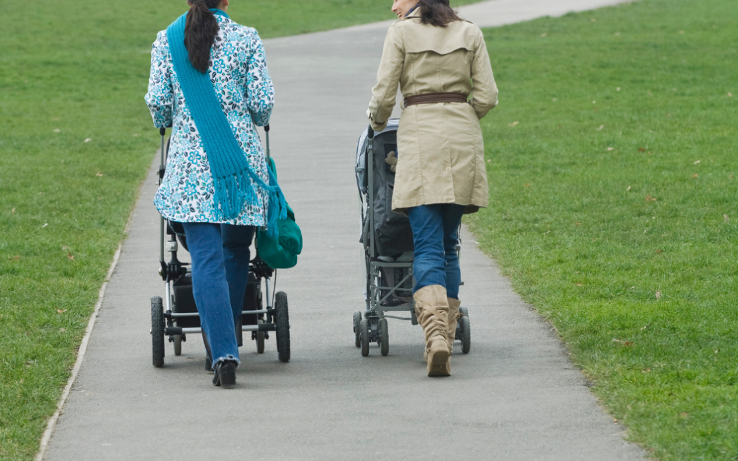 Two women walk side by side on a paved path in a grassy park, each pushing a stroller. The sky is overcast, and the path curves gently uphill in the distance.