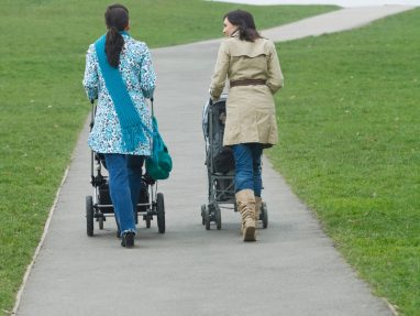 Two women walk side by side on a paved path in a grassy park, each pushing a stroller. The sky is overcast, and the path curves gently uphill in the distance.
