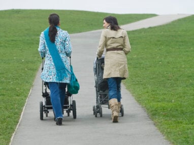 Two women walk side by side on a paved path in a grassy park, each pushing a stroller. The sky is overcast, and the path curves gently uphill in the distance.