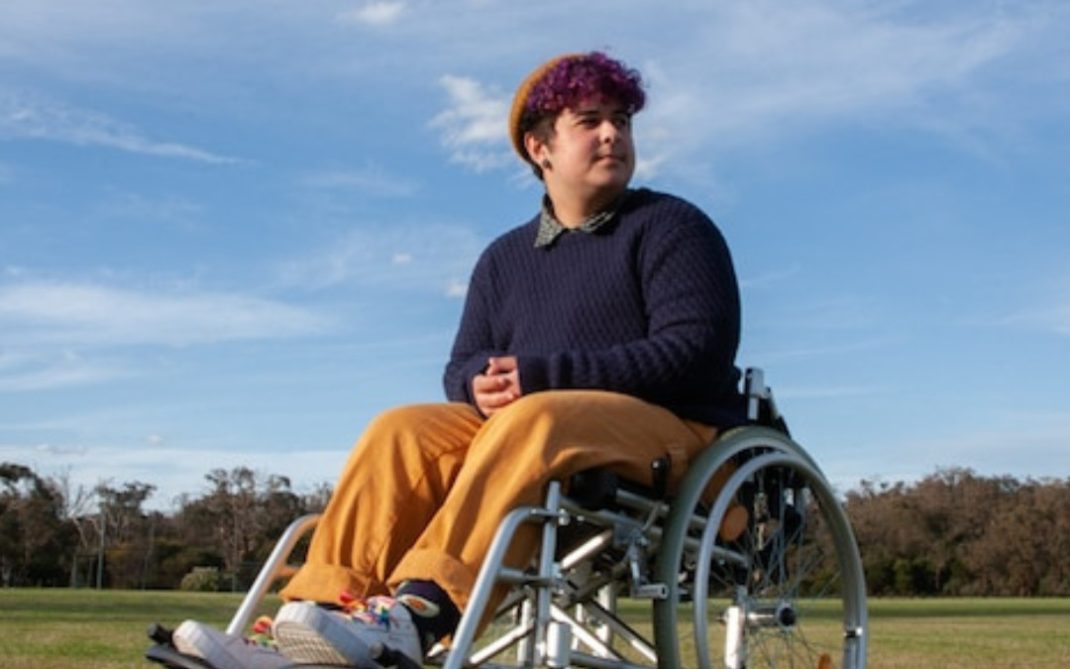 A person with short, curly purple hair sits in a wheelchair on grass under a blue sky, wearing a brown beanie, navy sweater, yellow pants, and sneakers, looking off into the distance.