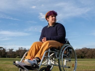 A person with short, curly purple hair sits in a wheelchair on grass under a blue sky, wearing a brown beanie, navy sweater, yellow pants, and sneakers, looking off into the distance.