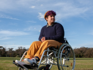 A person with short, curly purple hair sits in a wheelchair on grass under a blue sky, wearing a brown beanie, navy sweater, yellow pants, and sneakers, looking off into the distance.