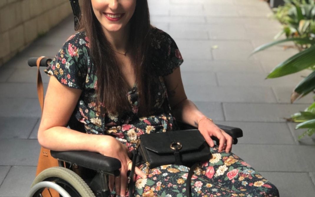 A woman with long brown hair, wearing a floral dress, sits in a wheelchair outdoors on a paved path lined with greenery. She is smiling and holding a black purse on her lap.