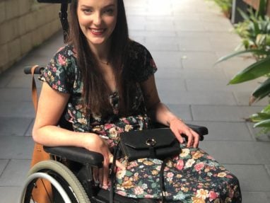 A woman with long brown hair, wearing a floral dress, sits in a wheelchair outdoors on a paved path lined with greenery. She is smiling and holding a black purse on her lap.