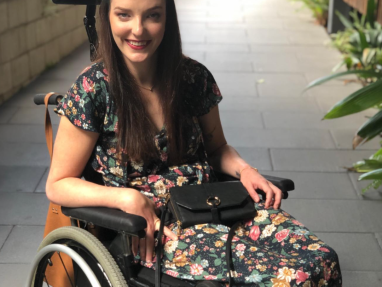 A woman with long brown hair, wearing a floral dress, sits in a wheelchair outdoors on a paved path lined with greenery. She is smiling and holding a black purse on her lap.