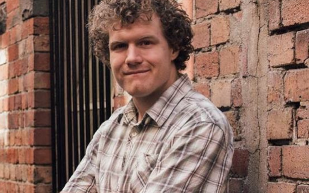 A person with curly hair and a plaid short-sleeve shirt stands with arms crossed, smiling, in front of a brick wall and window with bars.
