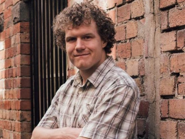 A person with curly hair and a plaid short-sleeve shirt stands with arms crossed, smiling, in front of a brick wall and window with bars.
