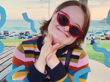 A smiling young woman wearing red cat-eye sunglasses and a colorful striped sweater poses with her hands clasped. Blue squiggles decorate the image, and a beach with parked cars is visible in the background.