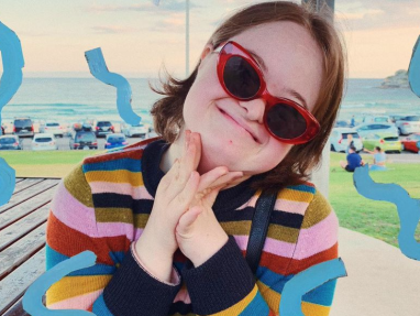 A smiling young woman wearing red cat-eye sunglasses and a colorful striped sweater poses with her hands clasped. Blue squiggles decorate the image, and a beach with parked cars is visible in the background.