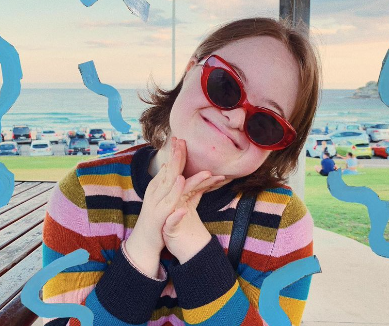A smiling young woman wearing red cat-eye sunglasses and a colorful striped sweater poses with her hands clasped. Blue squiggles decorate the image, and a beach with parked cars is visible in the background.
