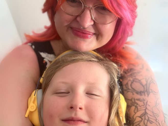 Parent with pink hair holding child wearing yellow headphones.