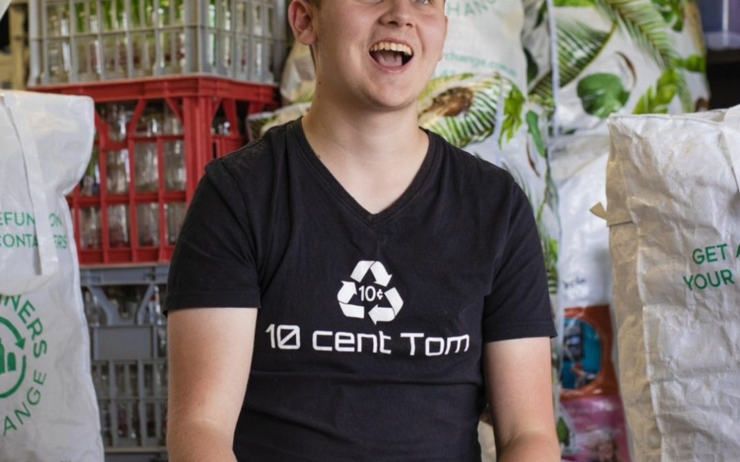 A smiling young man in a black “10 cent Tom” shirt sits in front of crates and bags filled with recyclable bottles and cans. The setting appears to be a recycling center or facility.