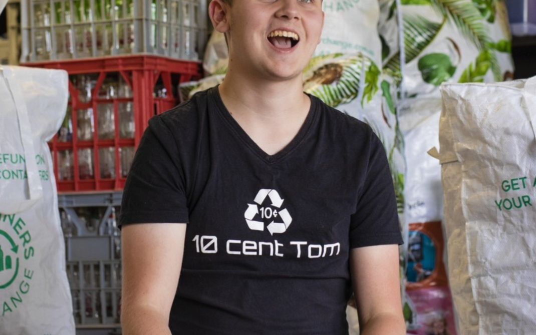 A smiling young man in a black “10 cent Tom” shirt sits in front of crates and bags filled with recyclable bottles and cans. The setting appears to be a recycling center or facility.
