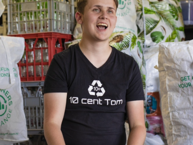 A smiling young man in a black “10 cent Tom” shirt sits in front of crates and bags filled with recyclable bottles and cans. The setting appears to be a recycling center or facility.