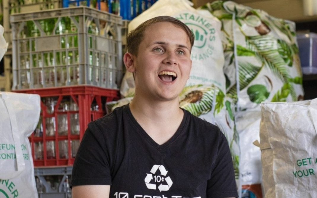 A smiling young man in a black “10 cent Tom” shirt sits in front of crates and bags filled with recyclable bottles and cans. The setting appears to be a recycling center or facility.
