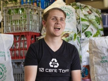 A smiling young man in a black “10 cent Tom” shirt sits in front of crates and bags filled with recyclable bottles and cans. The setting appears to be a recycling center or facility.