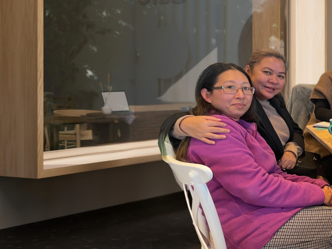 Six women sit together at a wooden table in a cafe, smiling at the camera. Cups, teapots, and small plates are on the table, and the background features a window and light-colored walls.