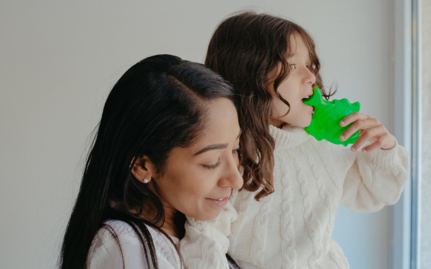 A woman with long dark hair smiles as she holds a young child with wavy brown hair who is wearing a white sweater and playing with a bright green toy near a window.