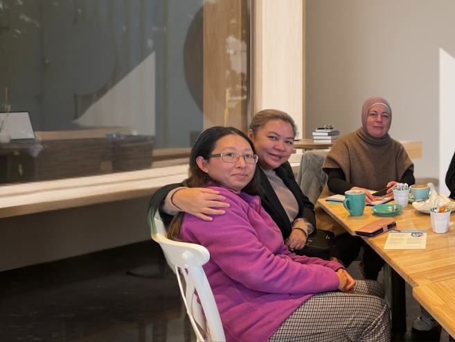 Six women sit together at a wooden table in a cafe, smiling at the camera. Cups, teapots, and small plates are on the table, and the background features a window and light-colored walls.