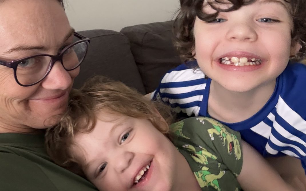 A mother sits on a couch with her two young sons close beside her, all looking toward the camera.
