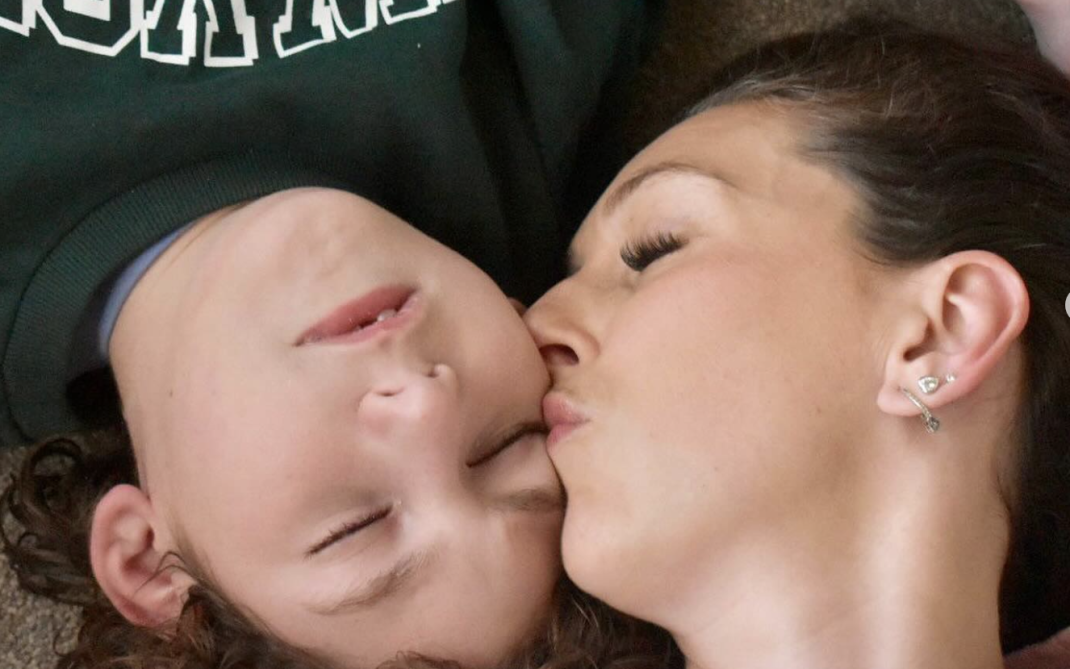 A mother lies beside her child with their heads touching, eyes closed, as she kisses the child’s cheek.