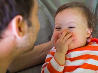 A close-up, heartwarming shot of a baby with Down syndrome laughing joyfully while looking at their father. The baby, wearing an orange and white striped shirt, has their hand playfully over their mouth. The father is seen from the back, slightly out of focus, as he gazes lovingly at the child.