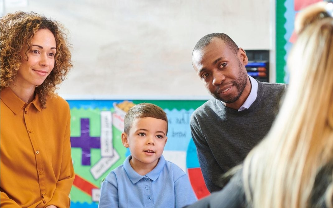 A family—a woman with curly brown hair, a young boy in a blue polo shirt, and a man in a grey sweater—sit together at a school table during a meeting. They are looking attentively toward an educator, whose blonde hair is visible in the foreground. The background features colorful classroom displays and educational posters, creating a professional yet supportive atmosphere.